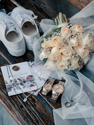 A wedding-themed arrangement with a pair of white sneakers adorned with lace, a bouquet of cream-colored roses with baby's breath, elegant invitations with a floral design and wax seal, two matching wristwatches, a pair of wedding rings, and delicate jewelry all placed on rustic wooden planks. Translucent white tulle drapes gracefully over several of these items, adding a touch of sophistication.