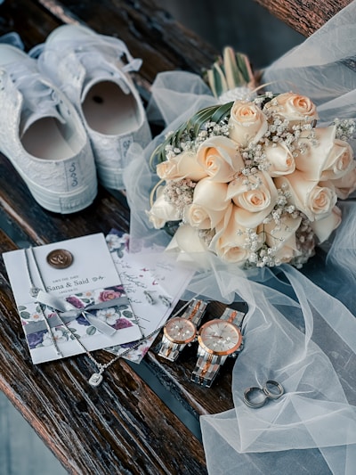 A wedding-themed arrangement with a pair of white sneakers adorned with lace, a bouquet of cream-colored roses with baby's breath, elegant invitations with a floral design and wax seal, two matching wristwatches, a pair of wedding rings, and delicate jewelry all placed on rustic wooden planks. Translucent white tulle drapes gracefully over several of these items, adding a touch of sophistication.