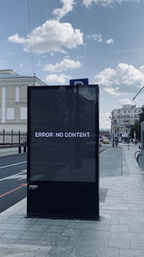 A large digital billboard on a city sidewalk displays the message 'ERROR: NO CONTENT'. The scene is set in an urban environment with buildings and street elements visible. The sky is partly cloudy, and the street is lined with vehicles.