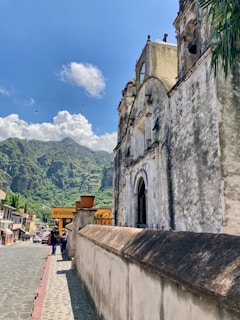 A historic stone building with arched windows and weathered textures is prominently featured. In the background, lush green mountains rise against a bright blue sky with scattered clouds. A cobblestone street leads past shops and pedestrians, adding a lively atmosphere to the scene. A sign reading 'Antigüedades' is visible along the street.