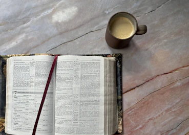 An open book with a striking crimson cover resting beside a steaming cup of coffee on a parchment-colored desk.