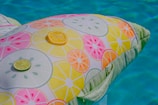 A colorful inflatable pool float is decorated with a pattern of citrus slices in yellow, orange, pink, and green. There are actual slices of lime and lemon placed on top of the float. The background features a clear, inviting blue pool.
