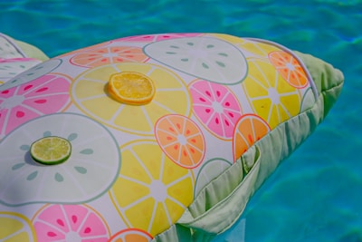 A colorful inflatable pool float is decorated with a pattern of citrus slices in yellow, orange, pink, and green. There are actual slices of lime and lemon placed on top of the float. The background features a clear, inviting blue pool.