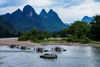 Several small boats with tourists are navigating a river, surrounded by lush green forests and dramatic limestone mountains. The sky is overcast, adding a serene and calm atmosphere to the scene.
