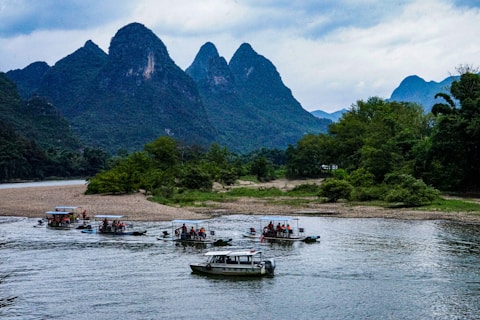 Several small boats with tourists are navigating a river, surrounded by lush green forests and dramatic limestone mountains. The sky is overcast, adding a serene and calm atmosphere to the scene.