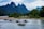 Several small boats with tourists are navigating a river, surrounded by lush green forests and dramatic limestone mountains. The sky is overcast, adding a serene and calm atmosphere to the scene.