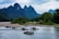 Several small boats with tourists are navigating a river, surrounded by lush green forests and dramatic limestone mountains. The sky is overcast, adding a serene and calm atmosphere to the scene.