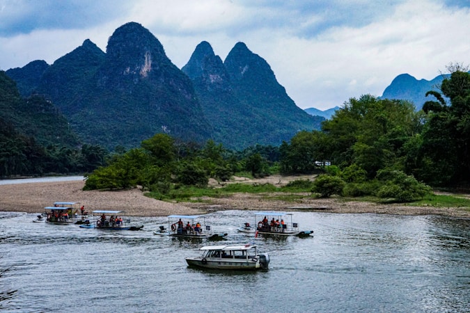 Several small boats with tourists are navigating a river, surrounded by lush green forests and dramatic limestone mountains. The sky is overcast, adding a serene and calm atmosphere to the scene.