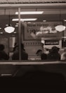 A dimly lit restaurant interior viewed through a window with people sitting at tables. A large sign in the background advertises job openings with a pay rate and a website for applications. Two hanging lights illuminate the area.