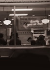A dimly lit restaurant interior viewed through a window with people sitting at tables. A large sign in the background advertises job openings with a pay rate and a website for applications. Two hanging lights illuminate the area.