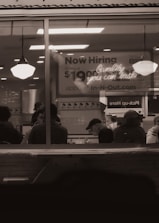 A dimly lit restaurant interior viewed through a window with people sitting at tables. A large sign in the background advertises job openings with a pay rate and a website for applications. Two hanging lights illuminate the area.