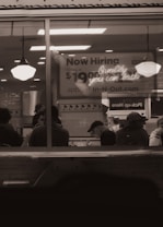 A dimly lit restaurant interior viewed through a window with people sitting at tables. A large sign in the background advertises job openings with a pay rate and a website for applications. Two hanging lights illuminate the area.
