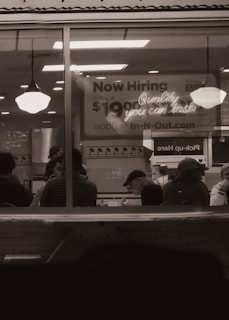 A dimly lit restaurant interior viewed through a window with people sitting at tables. A large sign in the background advertises job openings with a pay rate and a website for applications. Two hanging lights illuminate the area.