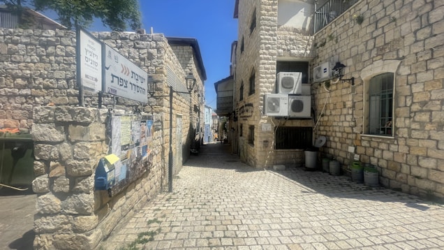 A narrow, cobblestone alleyway lined with stone buildings in a historic-looking area. Signs with Hebrew text are visible on the left, and there are outdoor air conditioning units mounted on the buildings. The alley leads to a brightly lit opening in the distance.