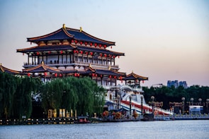 An intricately designed traditional Chinese building with multiple tiers and curved roofs adorned with red lanterns. Lush greenery surrounds the structure, which is situated beside a calm body of water. The skyline in the background includes modern buildings.