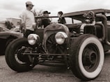 A vintage hot rod car is parked with its large front wheels prominently displayed. Three people stand nearby, interacting. The image has a sepia tone, enhancing the nostalgic feel.