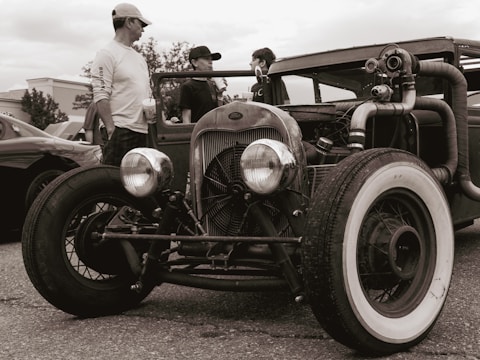 A vintage hot rod car is parked with its large front wheels prominently displayed. Three people stand nearby, interacting. The image has a sepia tone, enhancing the nostalgic feel.