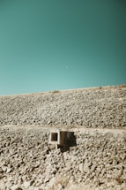 An expansive rocky landscape with a vivid teal sky backdrop. A small concrete structure, possibly a storm drain or culvert, sits among the stones, adding an element of human presence to the natural setting. The terrain is barren and rugged, stretching out to meet the horizon.