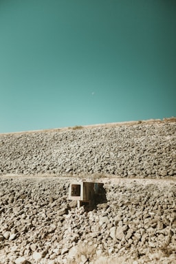 An expansive rocky landscape with a vivid teal sky backdrop. A small concrete structure, possibly a storm drain or culvert, sits among the stones, adding an element of human presence to the natural setting. The terrain is barren and rugged, stretching out to meet the horizon.