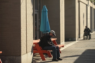 A person wearing a cap and backpack is sitting on an orange outdoor bench, looking at a device in their hands. A bright blue umbrella is placed near the bench. Another person in a wheelchair is visible farther down the paved pathway, near the edge of a building's shadow.