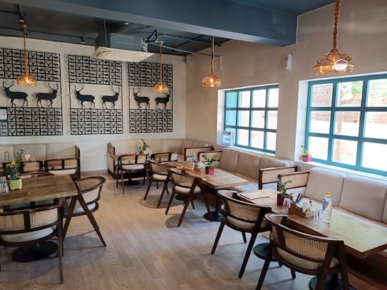 A cozy, well-decorated caf&eacute; featuring wooden tables and chairs with cushioned seating along the walls. The walls are adorned with artistic black deer silhouettes framed in checkered patterns. The space is well-lit with natural light coming through large blue-framed windows and warm hanging lamps. There are small plants placed on the tables and windowsills, adding a touch of greenery.