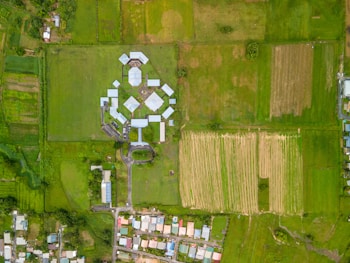 The aerial view features a rural landscape with extensive green fields and a cluster of buildings in a geometric pattern at the center. The buildings appear to be organized symmetrically, surrounded by lush green farmland. At the bottom of the image, a small residential area with colorful rooftops is visible, creating a contrast between developed and undeveloped land.