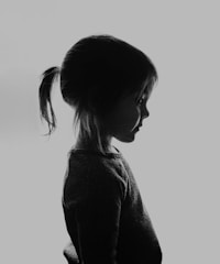 A silhouette of a young child with a ponytail is captured against a light background. The child is in profile, wearing a dark textured sweater, with the light emphasizing the contours of the face and hair.