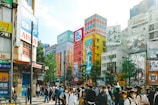 A busy urban street scene with numerous people, many of whom are wearing masks. Surrounding them are tall buildings adorned with vibrant advertisements, featuring anime characters and bold, colorful text. The scene conveys a bustling atmosphere with a mixture of pedestrians and urban signage.