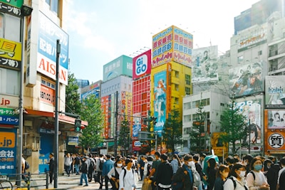 A busy urban street scene with numerous people, many of whom are wearing masks. Surrounding them are tall buildings adorned with vibrant advertisements, featuring anime characters and bold, colorful text. The scene conveys a bustling atmosphere with a mixture of pedestrians and urban signage.