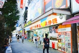 A bustling urban street scene featuring a variety of colorful shop signs and posters in Japanese. People walk along the sidewalk, some carrying shopping bags. A large, vibrant anime character billboard is visible above the store entrance. The shops display merchandise in the windows, attracting passersby.