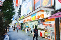 A bustling urban street scene featuring a variety of colorful shop signs and posters in Japanese. People walk along the sidewalk, some carrying shopping bags. A large, vibrant anime character billboard is visible above the store entrance. The shops display merchandise in the windows, attracting passersby.