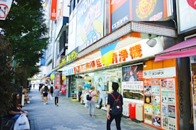 A bustling urban street scene featuring a variety of colorful shop signs and posters in Japanese. People walk along the sidewalk, some carrying shopping bags. A large, vibrant anime character billboard is visible above the store entrance. The shops display merchandise in the windows, attracting passersby.