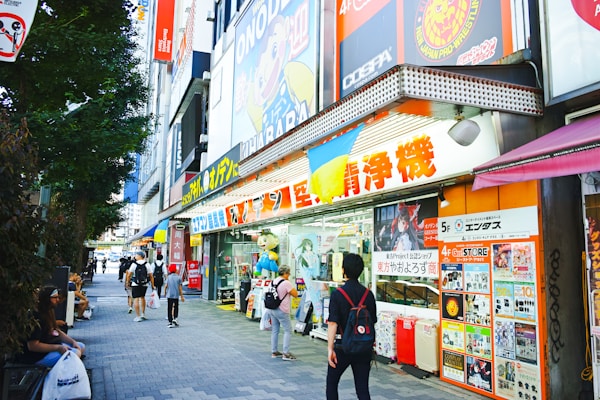 A bustling urban street scene featuring a variety of colorful shop signs and posters in Japanese. People walk along the sidewalk, some carrying shopping bags. A large, vibrant anime character billboard is visible above the store entrance. The shops display merchandise in the windows, attracting passersby.