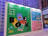 A vibrant advertisement board for the Hong Kong Book Fair 2023 is displayed. The board features colorful graphics including books, a ruler, and playful designs with large letters spelling 'BOOK FAIR'. The layout includes information about different exhibition halls and the World of Snacks event. The colors range from green, blue, to pink, creating an engaging atmosphere.