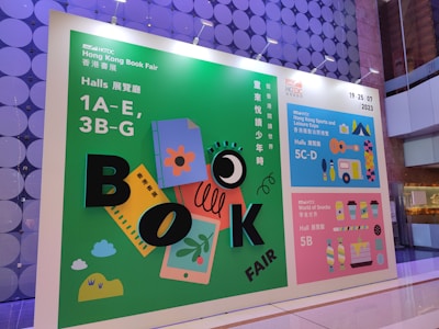 A vibrant advertisement board for the Hong Kong Book Fair 2023 is displayed. The board features colorful graphics including books, a ruler, and playful designs with large letters spelling 'BOOK FAIR'. The layout includes information about different exhibition halls and the World of Snacks event. The colors range from green, blue, to pink, creating an engaging atmosphere.