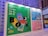 A vibrant advertisement board for the Hong Kong Book Fair 2023 is displayed. The board features colorful graphics including books, a ruler, and playful designs with large letters spelling 'BOOK FAIR'. The layout includes information about different exhibition halls and the World of Snacks event. The colors range from green, blue, to pink, creating an engaging atmosphere.