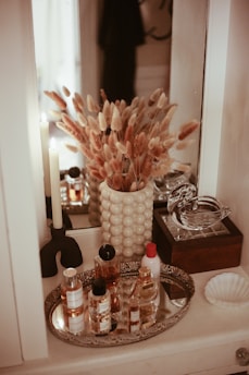 A mirror reflects a decorative arrangement of dried foliage in a textured vase, placed on a table. Nearby, a variety of skincare bottles are organized on a silver tray. A tall, elegant candle with a black holder stands beside the vase. A small, ornate box with a glass lid sits next to a shell-shaped dish, adding a touch of elegance to the scene.