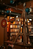 A miniature model resembling the Eiffel Tower sits atop a stack of books on a shelf in a warmly lit library. Suspended around it are various circular objects, including magnifying glasses and blue spheres, suggesting a whimsical and creative atmosphere. In the background, bookshelves filled with various books add depth and richness to the scene.