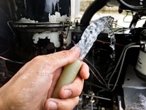 A hand is holding a water hose with clear water flowing out against the backdrop of a mechanical or industrial setting, including pipes and machinery.