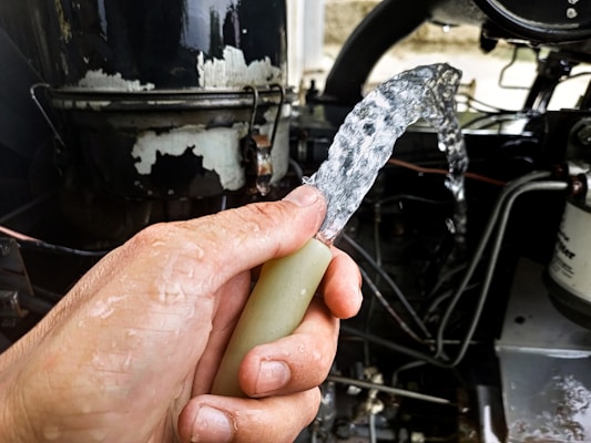 A hand is holding a water hose with clear water flowing out against the backdrop of a mechanical or industrial setting, including pipes and machinery.