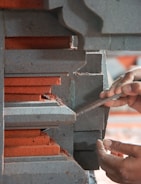 A person's hands are using a tool to work on a piece of layered concrete or stone structure with red and gray colors. The layers have intricate designs and appear to be part of a larger architectural element.