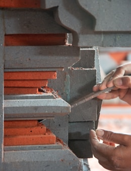 A person's hands are using a tool to work on a piece of layered concrete or stone structure with red and gray colors. The layers have intricate designs and appear to be part of a larger architectural element.