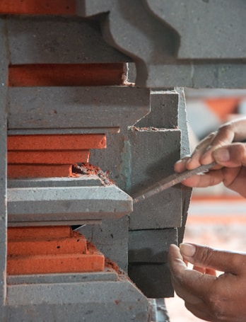 A person's hands are using a tool to work on a piece of layered concrete or stone structure with red and gray colors. The layers have intricate designs and appear to be part of a larger architectural element.