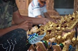 A person is painting a detailed sculpture with intricate designs, featuring gold, red, and blue colors. The sculpture appears to be a decorative piece with ornate patterns and a figure with wings.