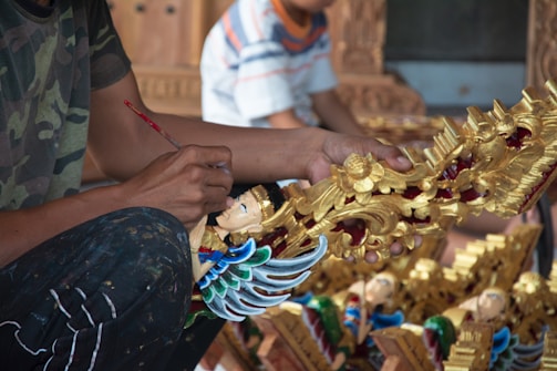 A person is painting a detailed sculpture with intricate designs, featuring gold, red, and blue colors. The sculpture appears to be a decorative piece with ornate patterns and a figure with wings.