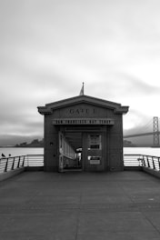 A black and white image featuring the entrance of Gate E for the San Francisco Bay Ferry. The structure is framed symmetrically, with a bridge partially visible in the background to the right. The scene includes a wide walkway leading up to the gate, with railings on either side.