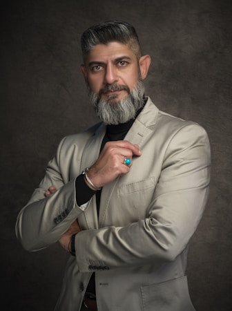 A man with a well-groomed beard and mustache is wearing a light gray suit over a black shirt. He stands against a textured dark backdrop with a serious and confident expression. His arms are folded, and he is holding his chin partially with one hand, which is adorned with a turquoise ring and a bracelet.