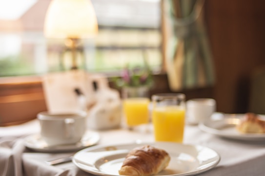 A breakfast table setup with a croissant on a plate in the foreground, a cup of coffee and glasses of orange juice are on the table. The background includes a lamp and a window with natural light coming through. The scene suggests a cozy atmosphere.