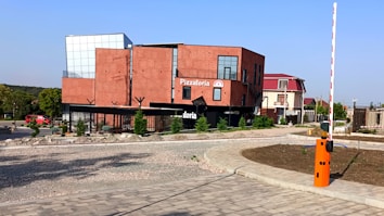 A modern, rectangular red-brick building with black accents serves as a pizzeria, labeled 'Pizzatoria'. It is situated on a corner next to a paved driveway and surrounded by a few small trees. The building features large glass windows and a flat rooftop with metal railings. Nearby, a red vehicle is parked along a tree-lined street. A barrier gate with an orange control panel is visible in the foreground, indicating controlled access.