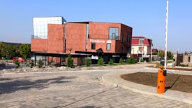 A modern, rectangular red-brick building with black accents serves as a pizzeria, labeled 'Pizzatoria'. It is situated on a corner next to a paved driveway and surrounded by a few small trees. The building features large glass windows and a flat rooftop with metal railings. Nearby, a red vehicle is parked along a tree-lined street. A barrier gate with an orange control panel is visible in the foreground, indicating controlled access.